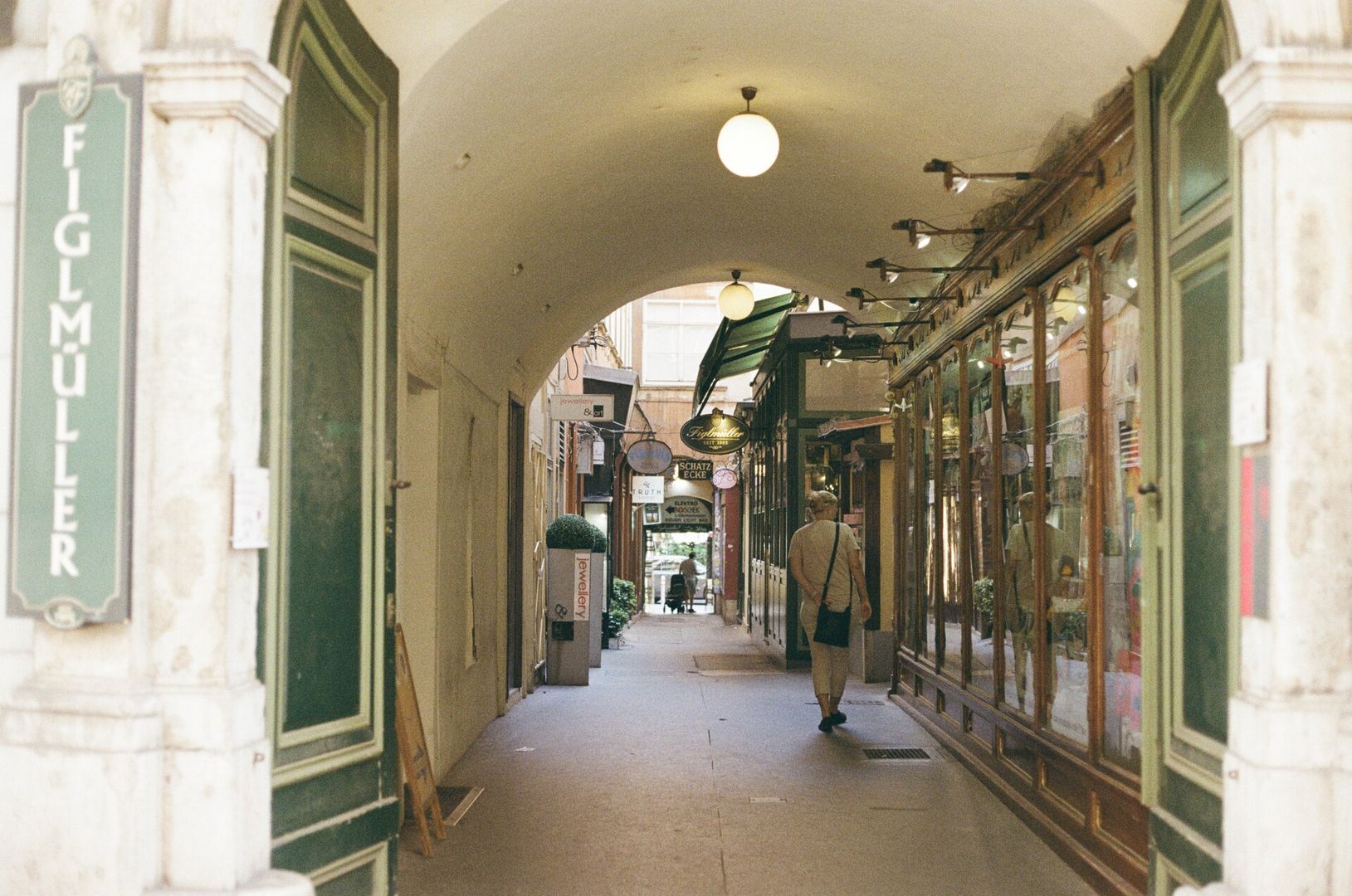 A covered passage through a building, seen along its length. The vaulted ceiling curves overhead and a round white globe lamp hangs from it in the foreground, with a second one further back. On the left is a heavy stone pillar and a tall green-painted shopfront with a vertical sign reading FIGLMÜLLER. On the right runs a row of old wooden and glass shop windows with brass fittings and small lamps. In the middle distance a person in light clothing with a shoulder bag walks away from the camera along the stone floor. Beyond them the passage opens into daylight, with hanging shop signs including SCHATZ ECKE. Colour photograph on 35mm film, with visible grain.